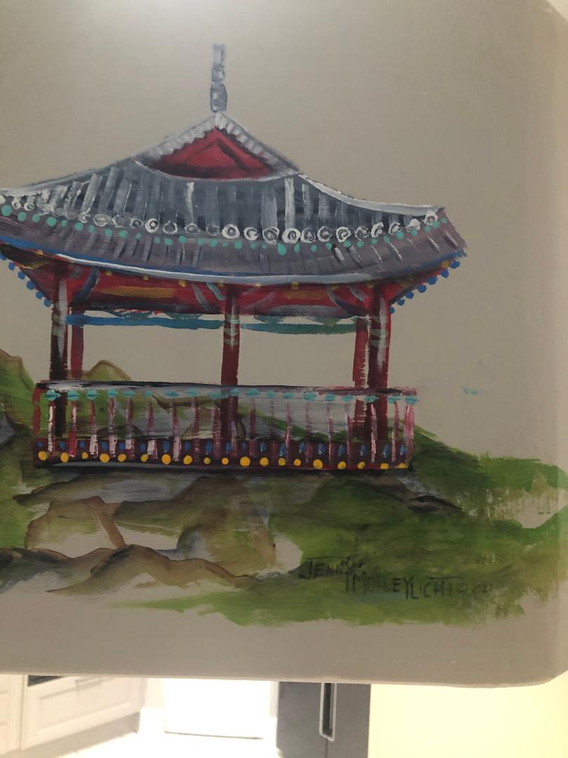 A tiled pavilion with an upswept red and grey roof painted on a wall corner above a green hillside.