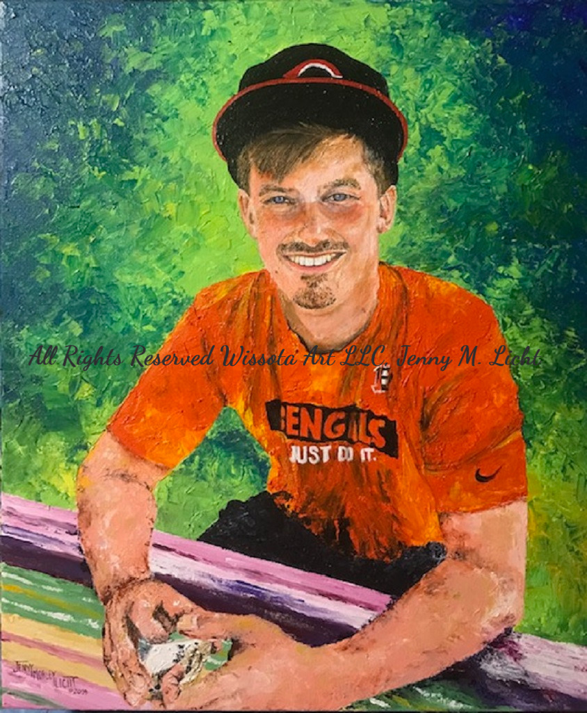 A young man in an orange t-shirt and ball cap leaning on a brightly striped railing, green foliage behind him.