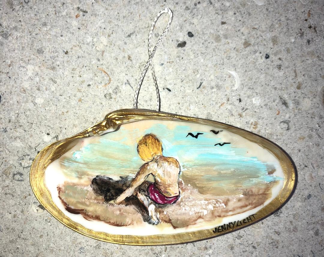 A gilt-edged shell ornament painted with a child sitting at the water's edge beside a dog, gulls in the distance.