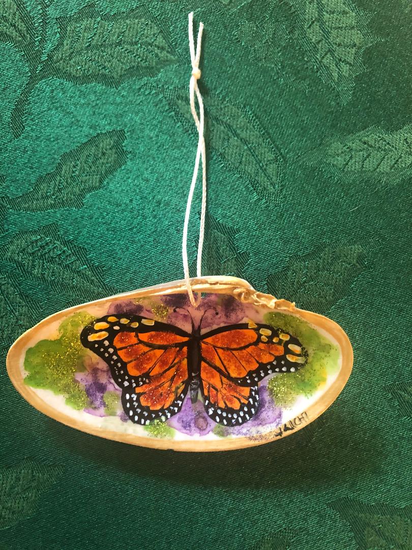 A monarch butterfly painted inside a polished shell, hung on a cotton loop, with glitter-flecked green and violet around it.