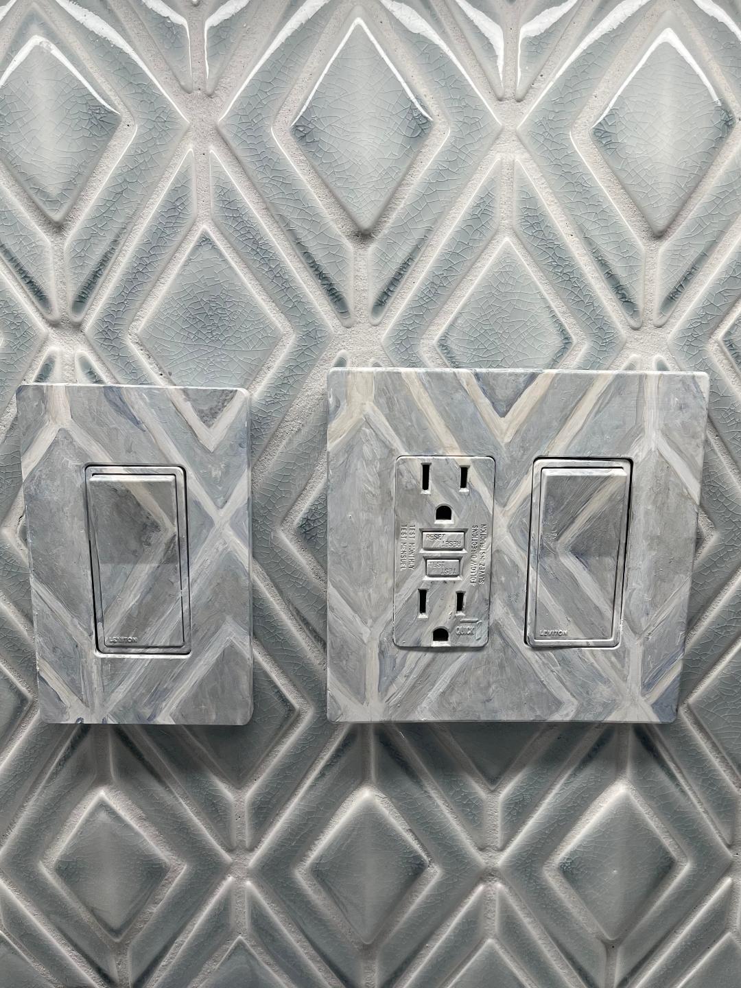 A switch plate and a combination outlet plate painted to match the pale grey diamond tile they sit on.